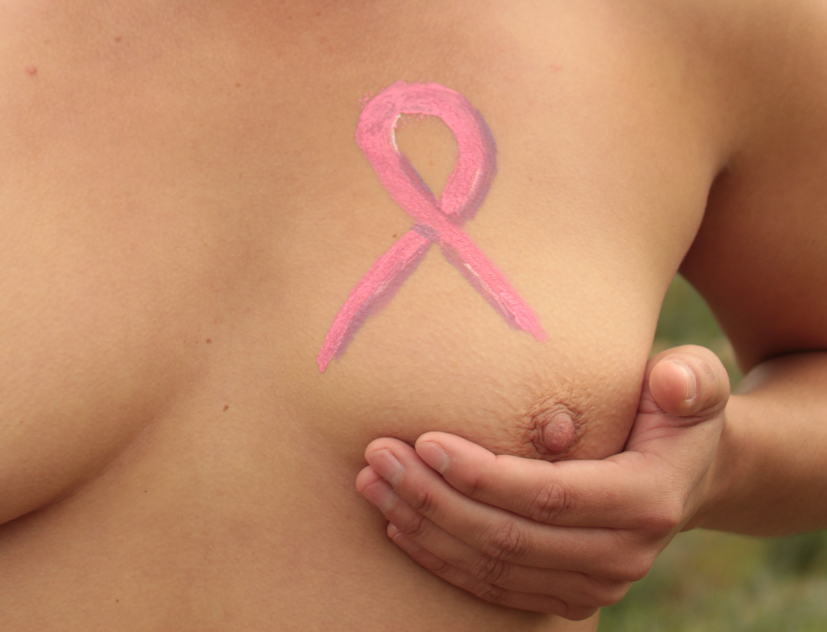 topless woman with pink ribbon body painted on her left breast; she cups that breast in her left hand.