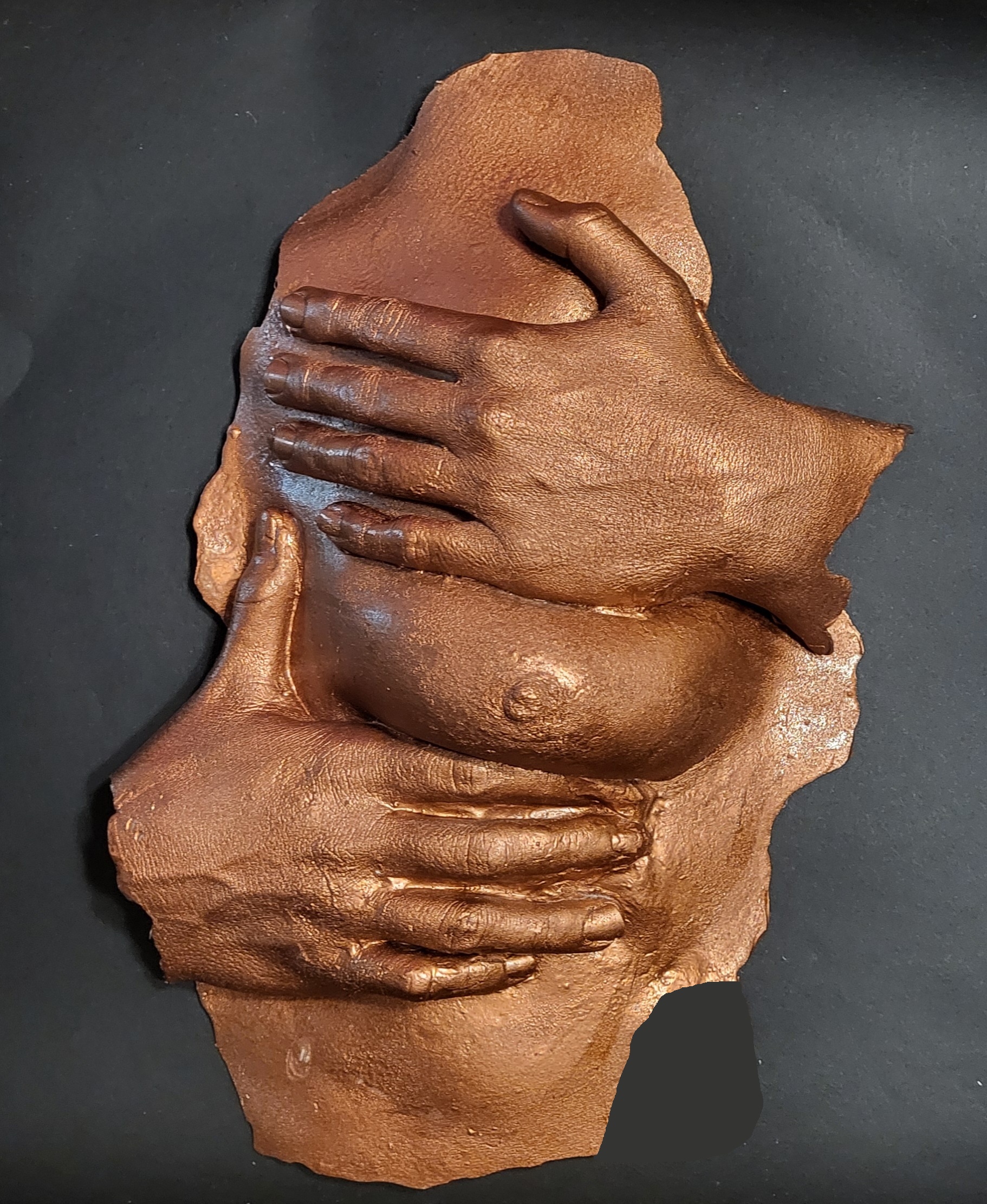 white plastic casting, painted in copper color, of woman's left breast with her left hand above it and right hand below it
